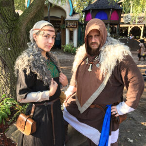 A couple dressed in medieval garments at a Renaissance Faire, standing by a tree, with the man wearing a hood and the woman a head scarf.