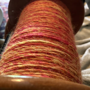 Close-up view of a wooden spindle tightly wound with handspun yarn in shades of orange and red.