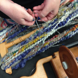 Hands weaving multicolored wool on a wooden loom.