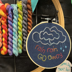 A craft display featuring colorful yarns and a blue fabric embroidery hoop with the phrase "rain rain GO away" stitched in.