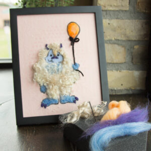 A whimsical needle felted yeti holding an orange balloon, displayed in a framed artwork setting.