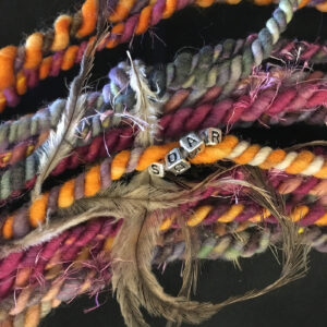 A close-up view of colorful handspun yarn with embedded letters spelling "SOAR" and adorned with feathers.