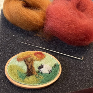 A needle felting landscape showing a whimsical scene with a tree and a sheep, displayed in a small wooden hoop.